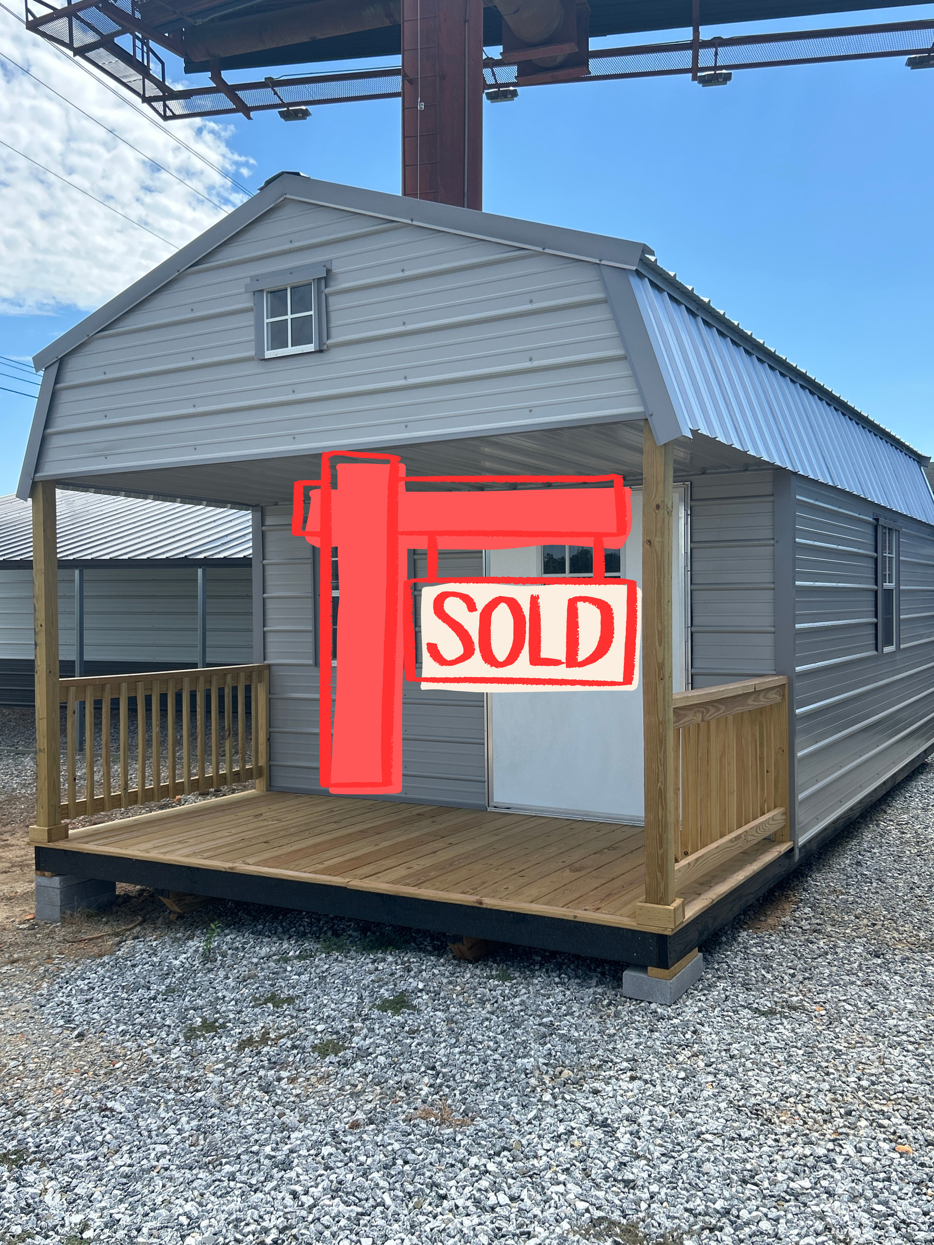 Gray cabin with porch, gray siding, white door, blue roof, set on gravel.