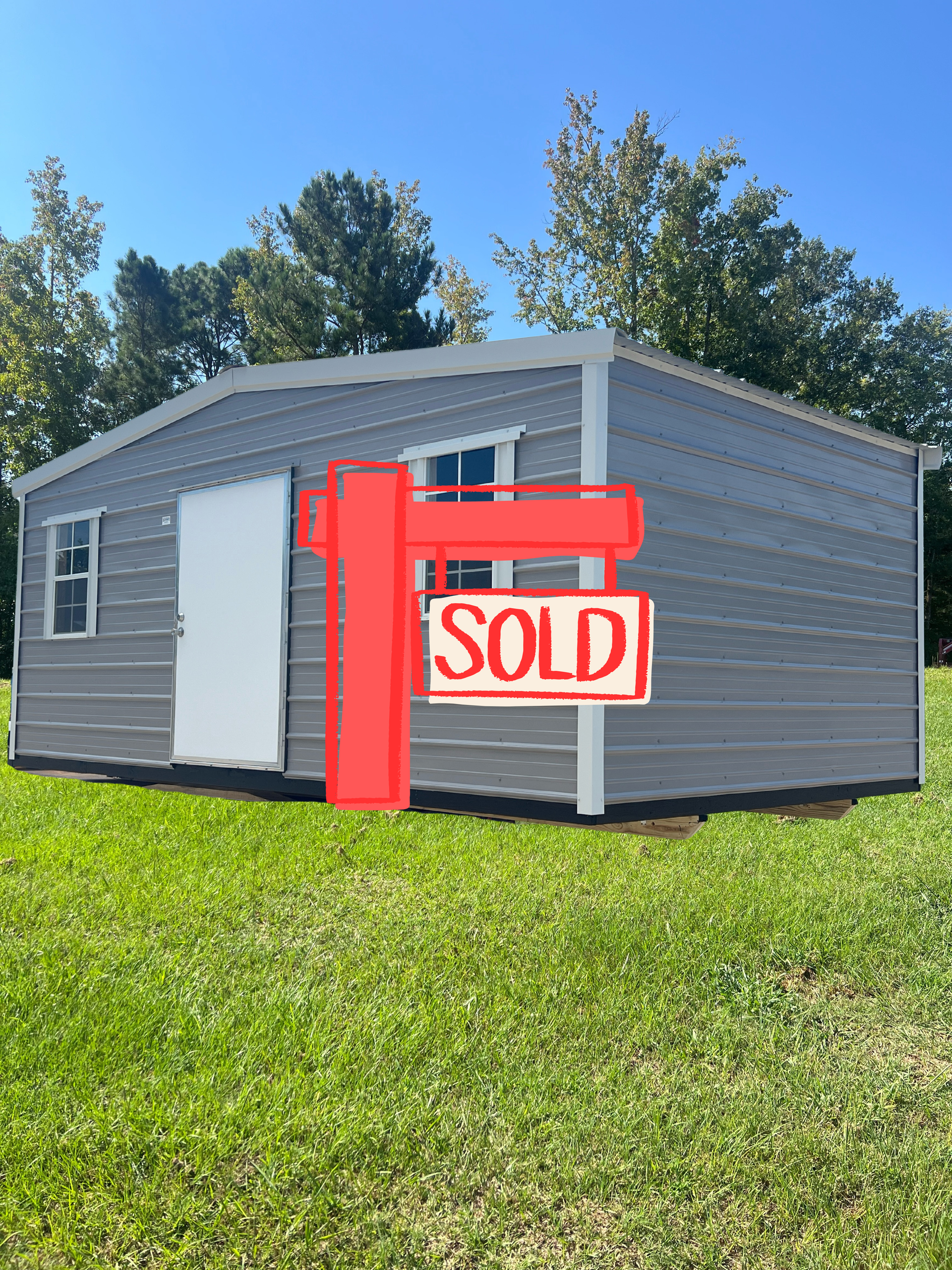 Gray shed on a grassy lot with a red “SOLD” sign in front under a blue sky