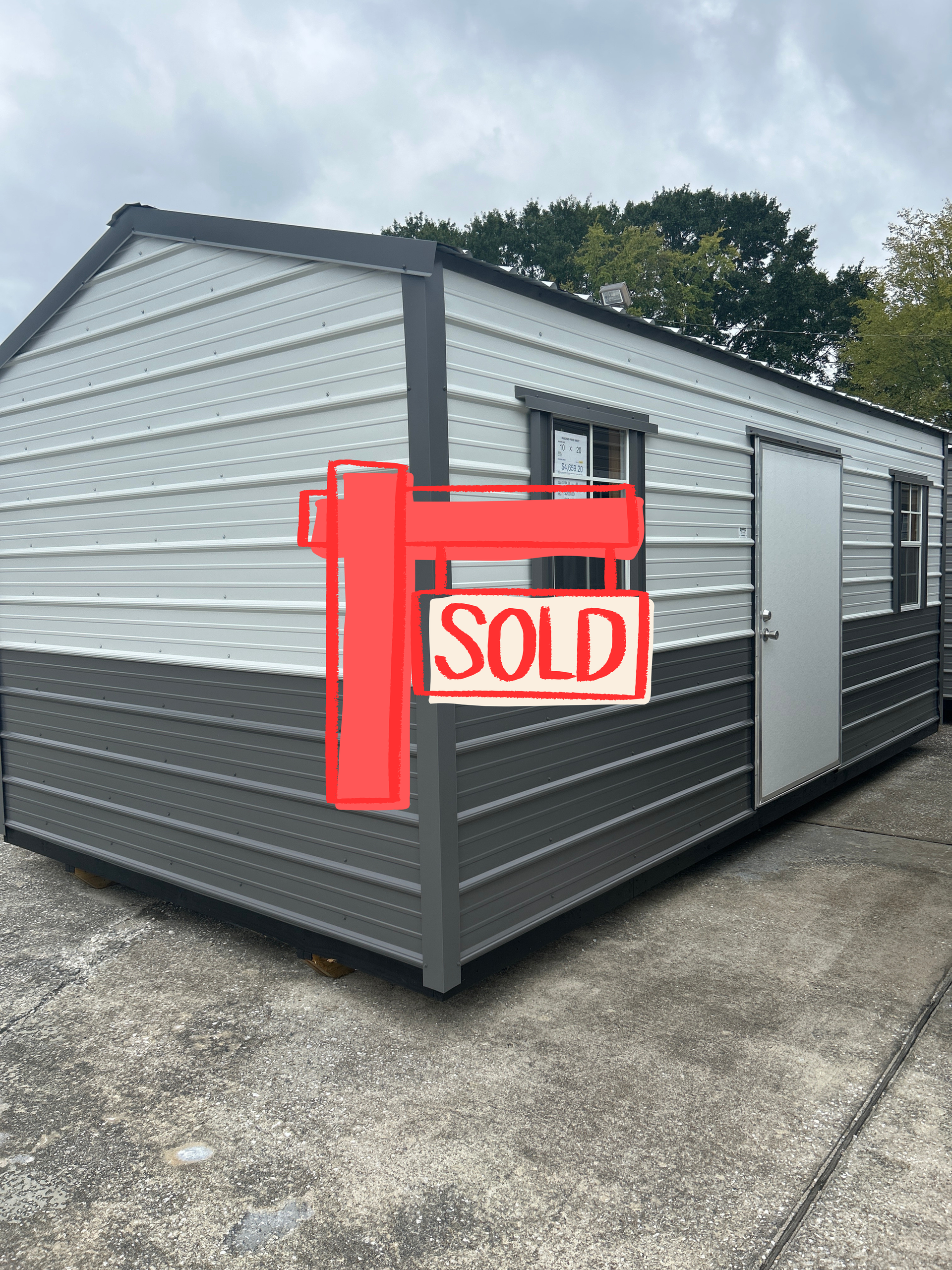 White and gray portable building with a red SOLD sign on the side, parked on a concrete lot.