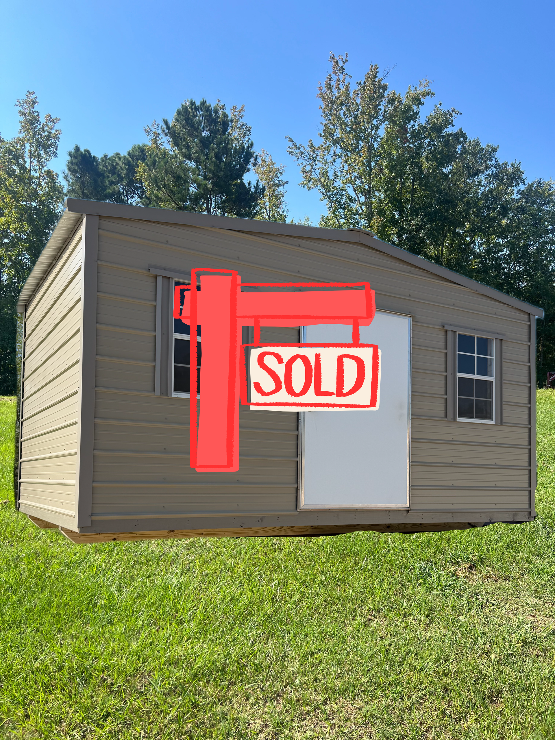 Small beige shed on grass with a red “SOLD” sign on the front and trees in the background