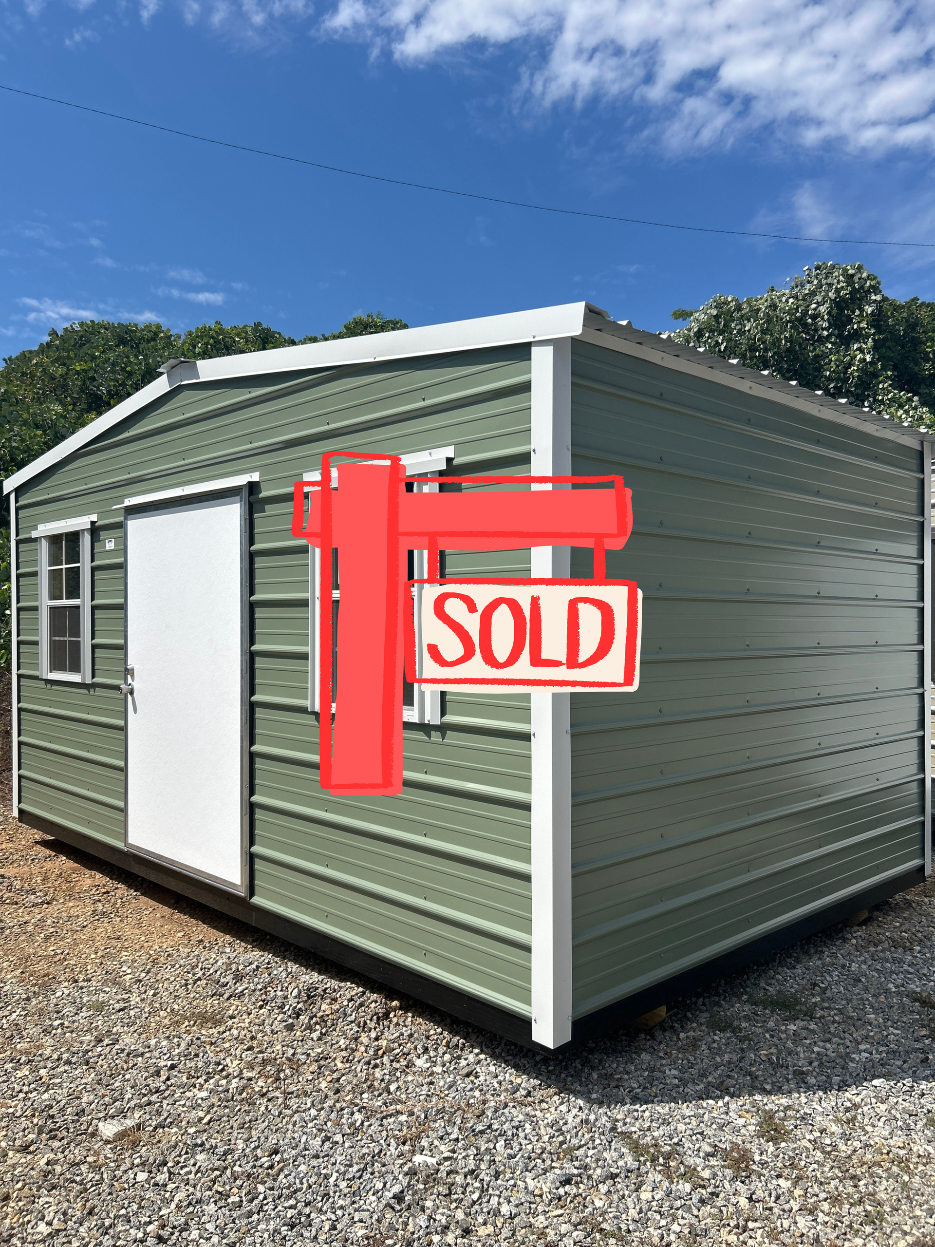 Green metal storage shed with white trim, door, and windows. Outdoors, gravel ground, blue sky.