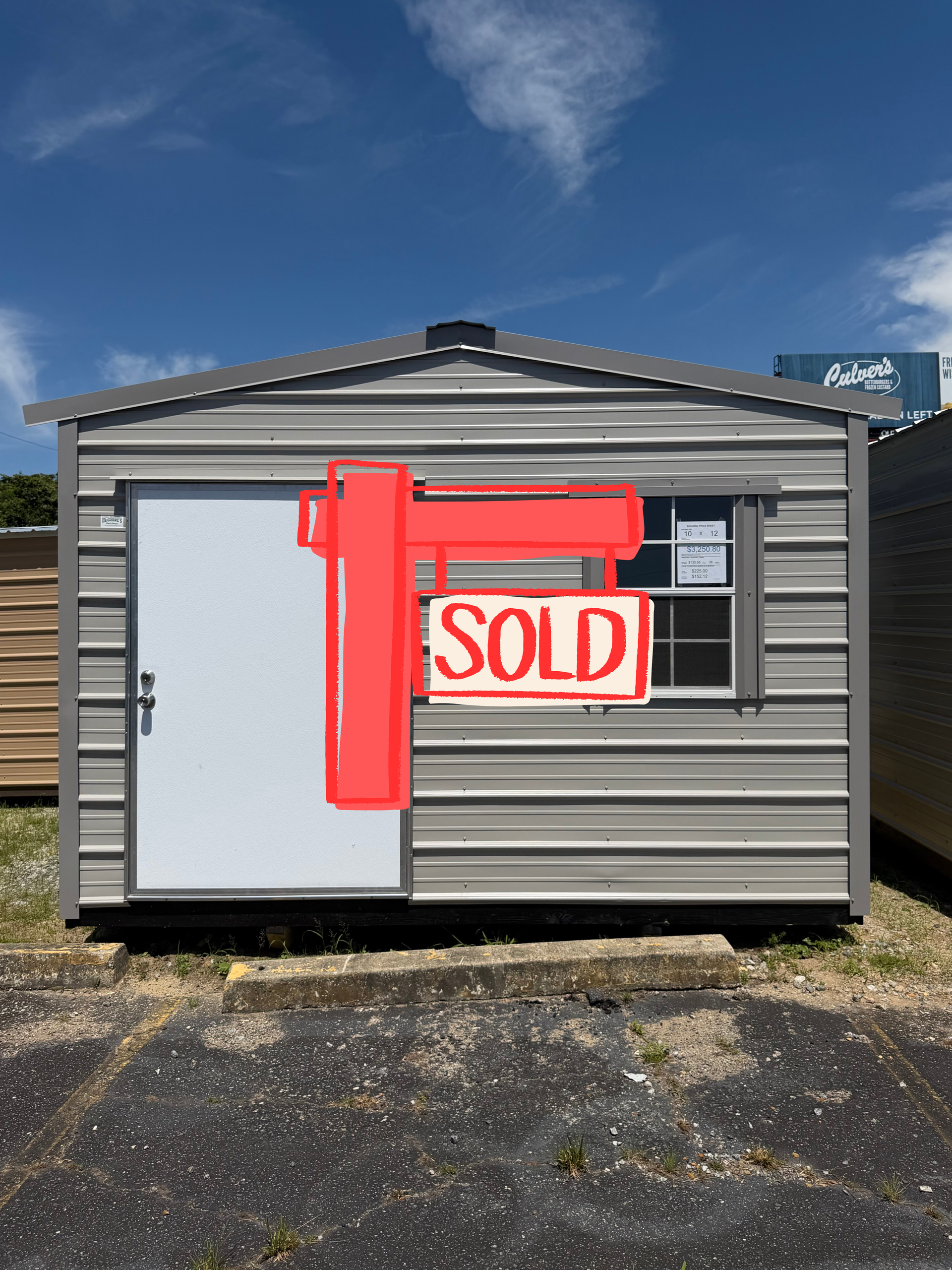 Small gray mobile home with a red “SOLD” sign on the front, set on a gravel lot under a blue sky.