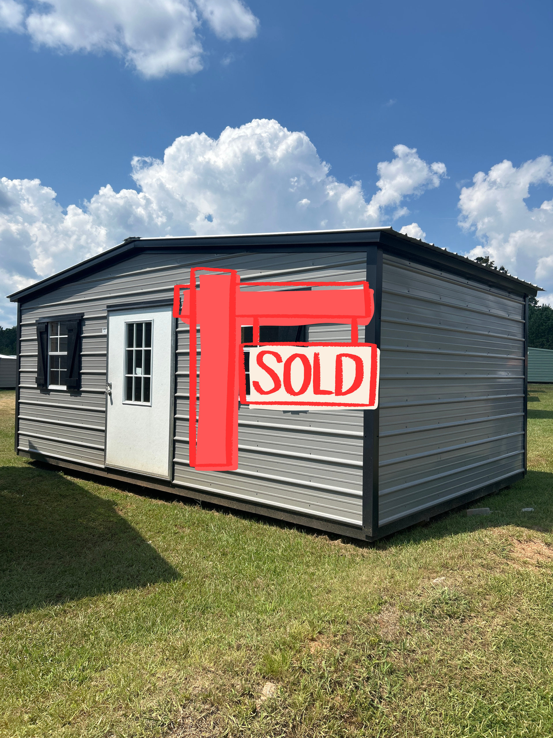 Gray shed with black trim, white door, black shutters, and a cloudy blue sky.
