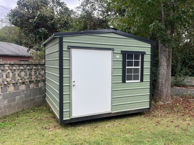Green metal shed with white door, black trim, and small window in a yard.
