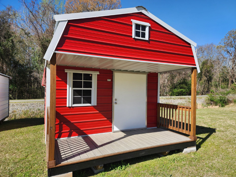 Red barn-style shed with porch and white trim on grass under a clear blue sky.