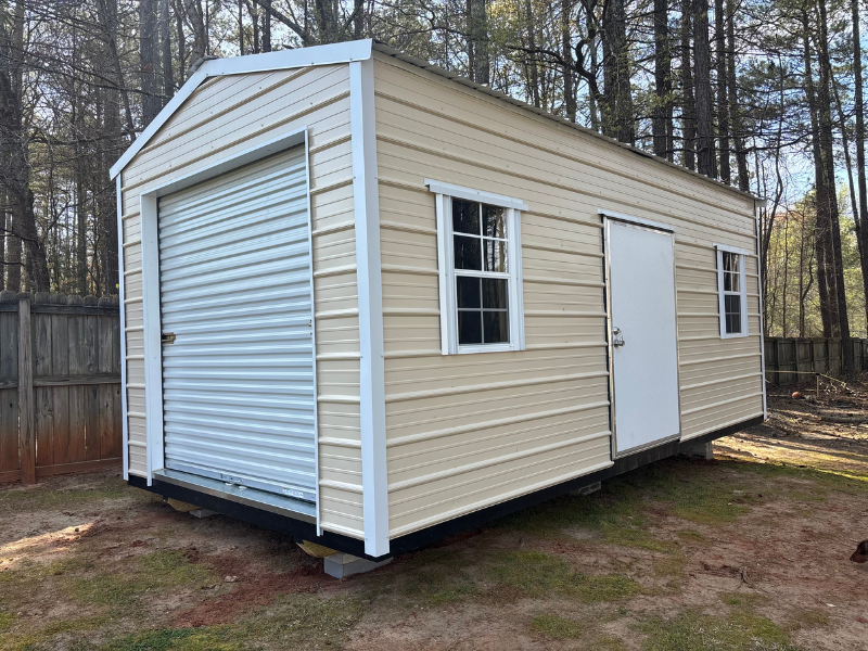 Tan metal shed with a roll-up garage door, a white door, and two windows.