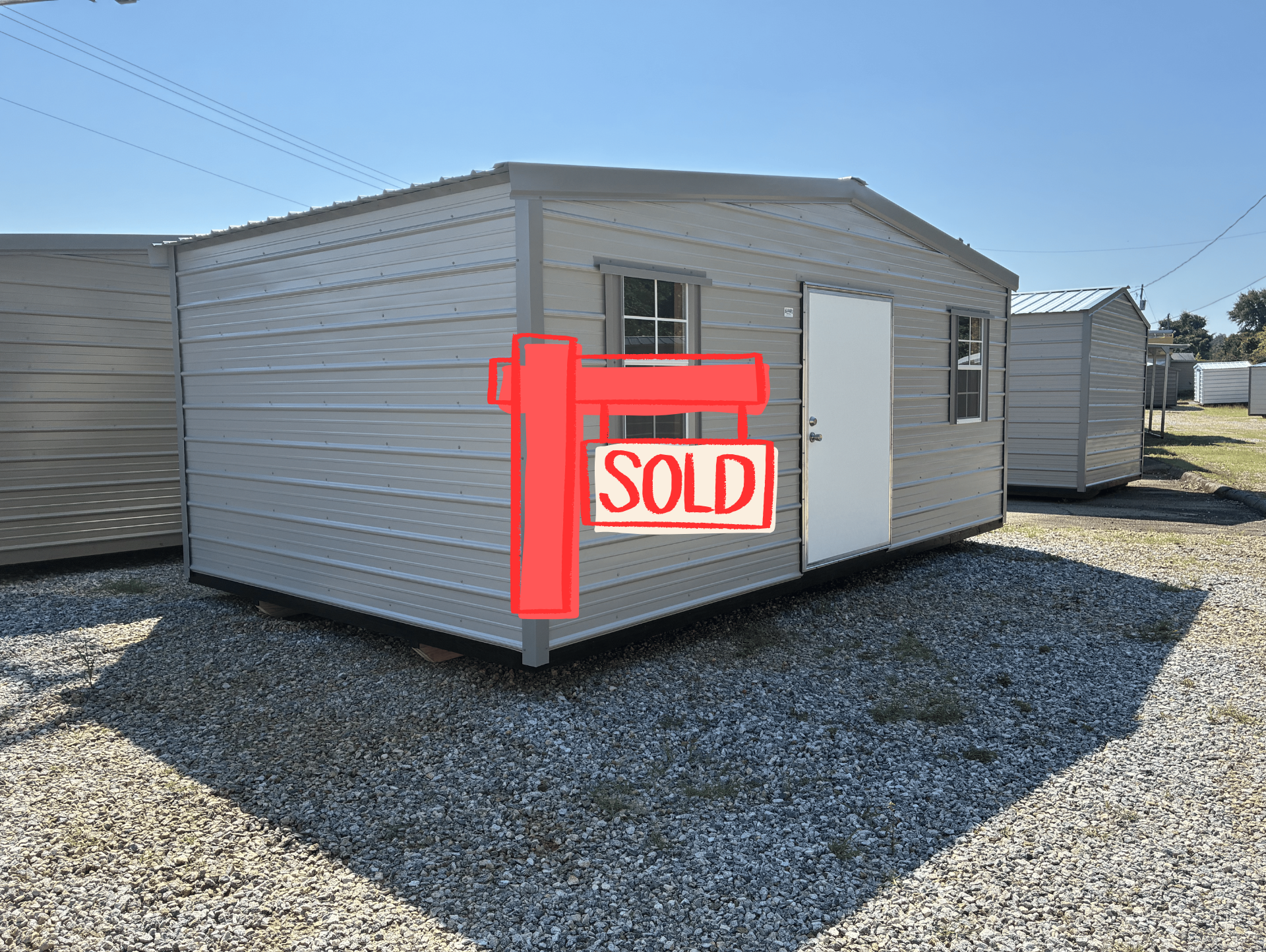 Small gray shed with windows and a white door on a gravel lot under a clear sky
