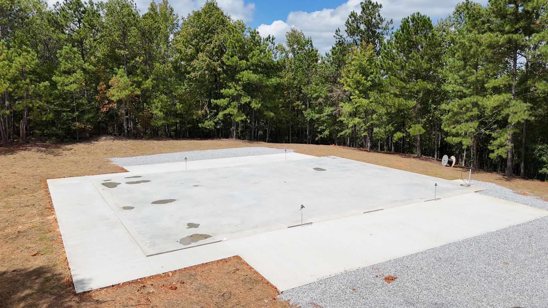 Concrete foundation, surrounded by gravel and grass, in a wooded area under a blue sky.