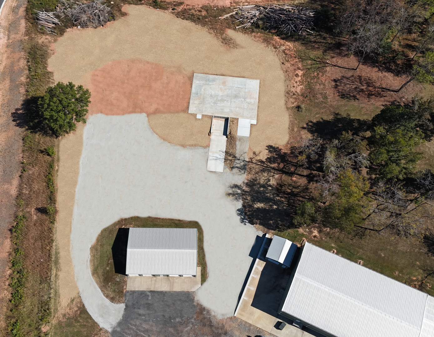 Aerial view of a gravel yard with a covered area, a rectangular building, and several trees.