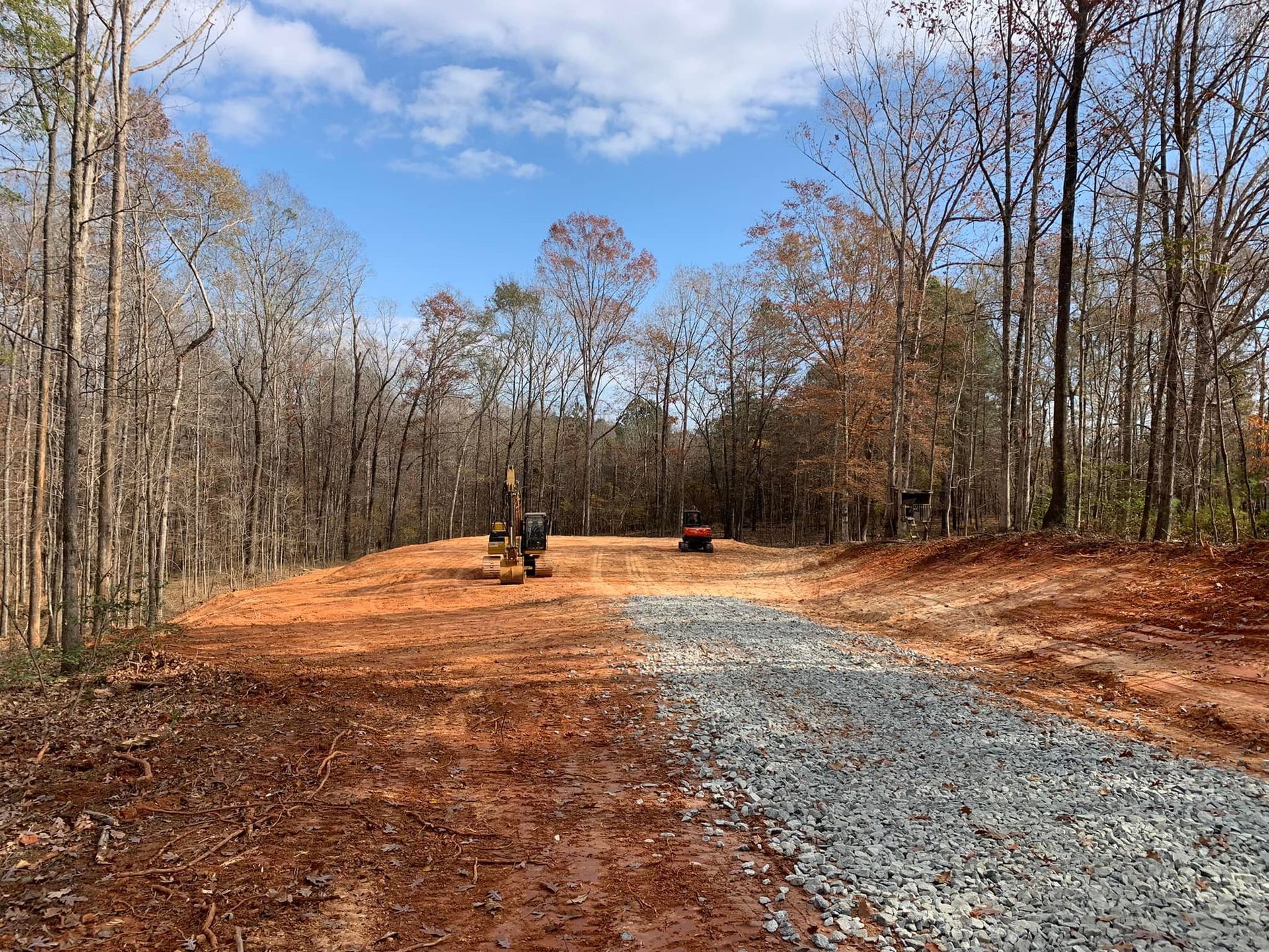 Clearing in woods with gravel path, red dirt, and construction equipment under a partly cloudy sky.