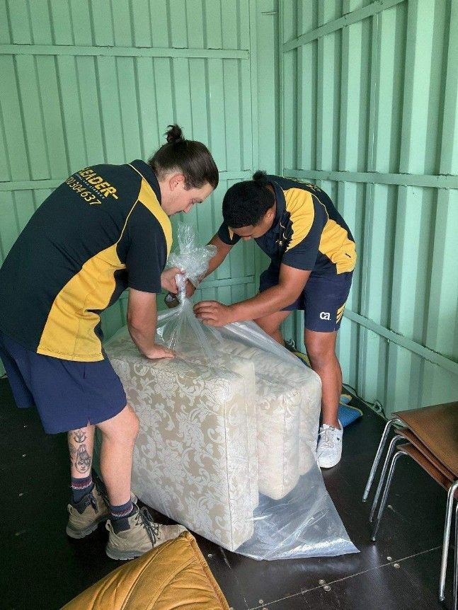 Two movers wrapping a beige couch in plastic inside a storage unit -  — Leader Removals & Storage in Mitchell, ACT