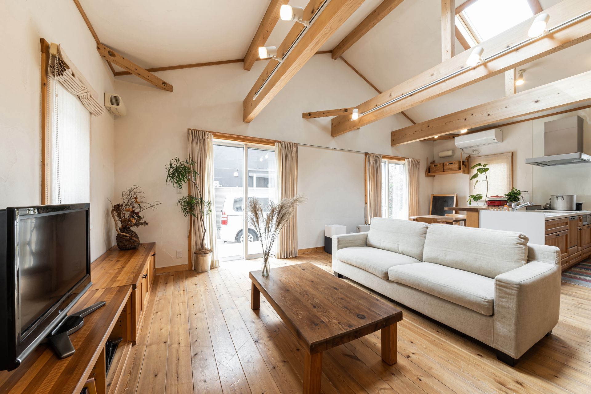 Spacious living room with wooden beams, sofa, TV, and kitchen in the background. Natural light floods the space.