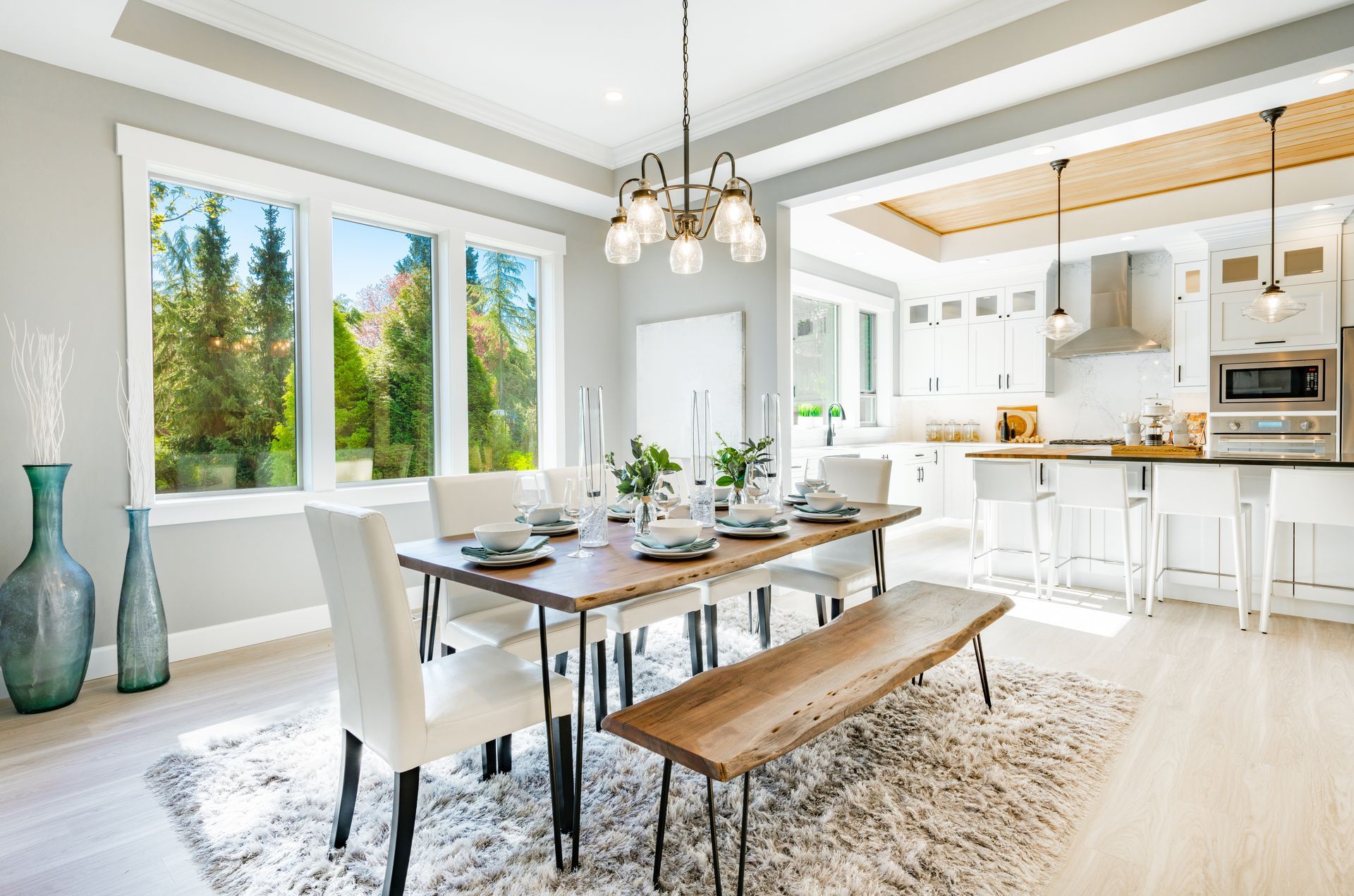 Dining room with a wooden table, white chairs, and large windows overlooking trees.