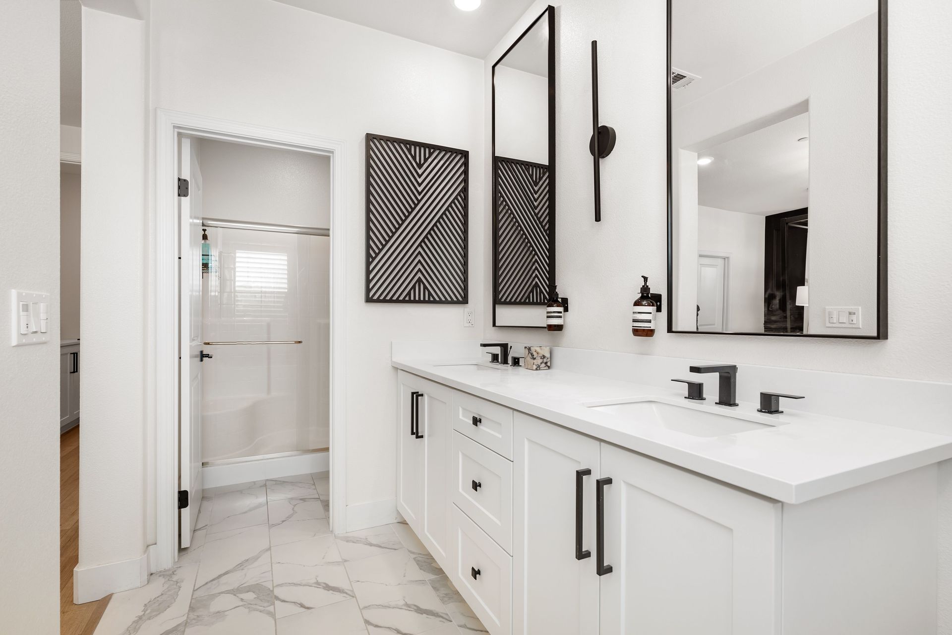 White bathroom with dual sinks, black fixtures, and a shower in the back.