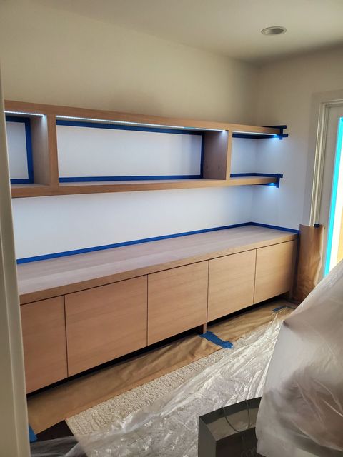 Built-in wooden shelving unit with cabinets, shelves, and recessed lighting against a white wall; blue tape lines.