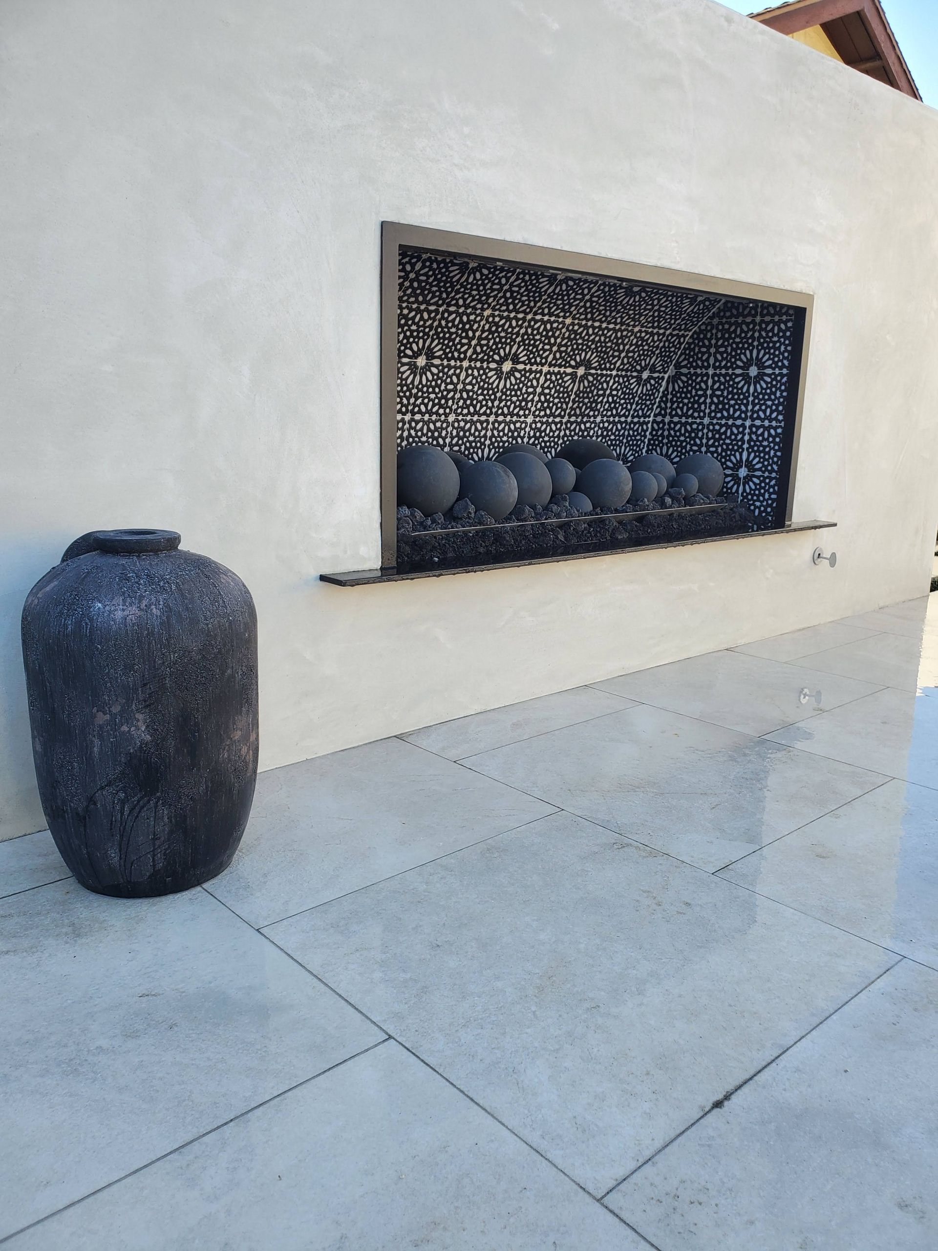 Large gray vase next to a white stucco wall with a window displaying dark pots.