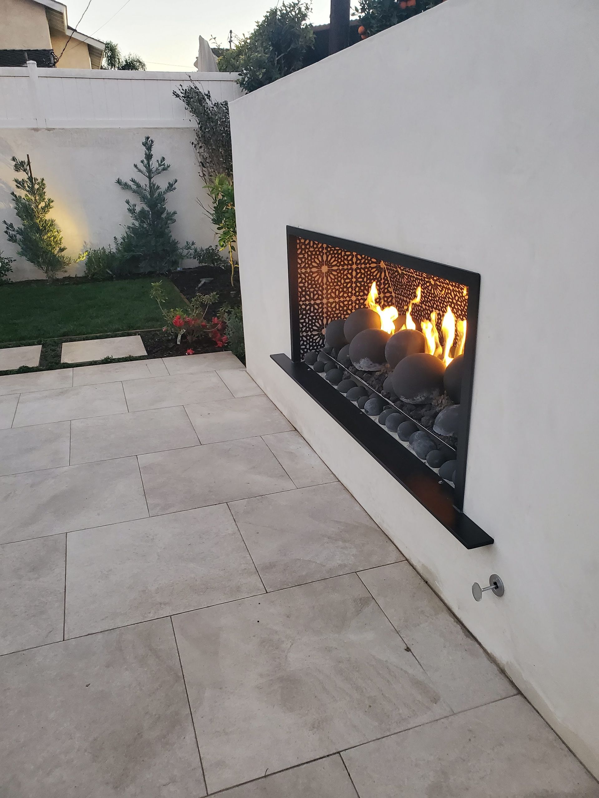 Outdoor fireplace built into a white wall, flames visible behind a decorative screen, patio.