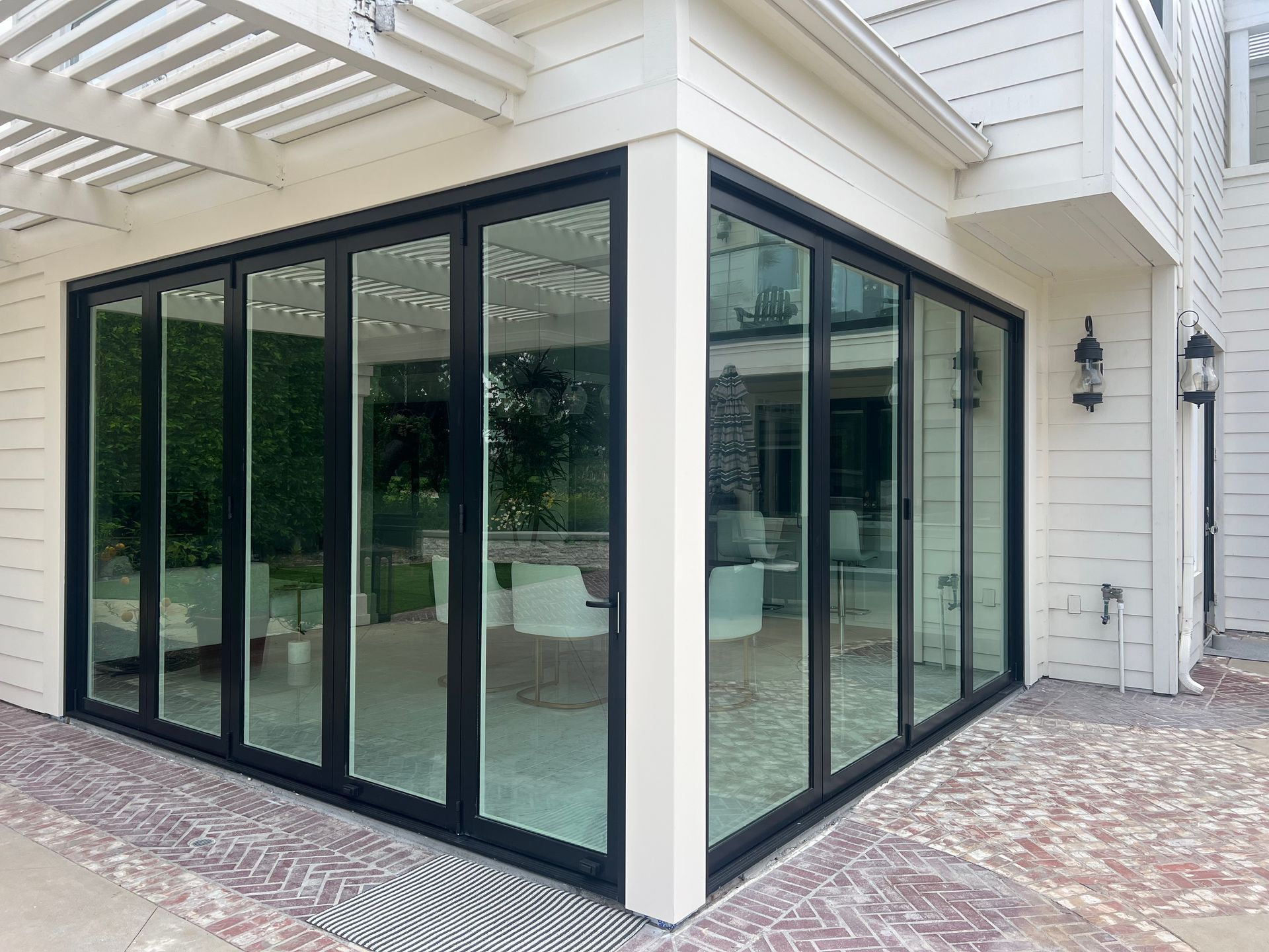 Black-framed glass folding doors on a white building corner, opening to a patio with brick pavers.