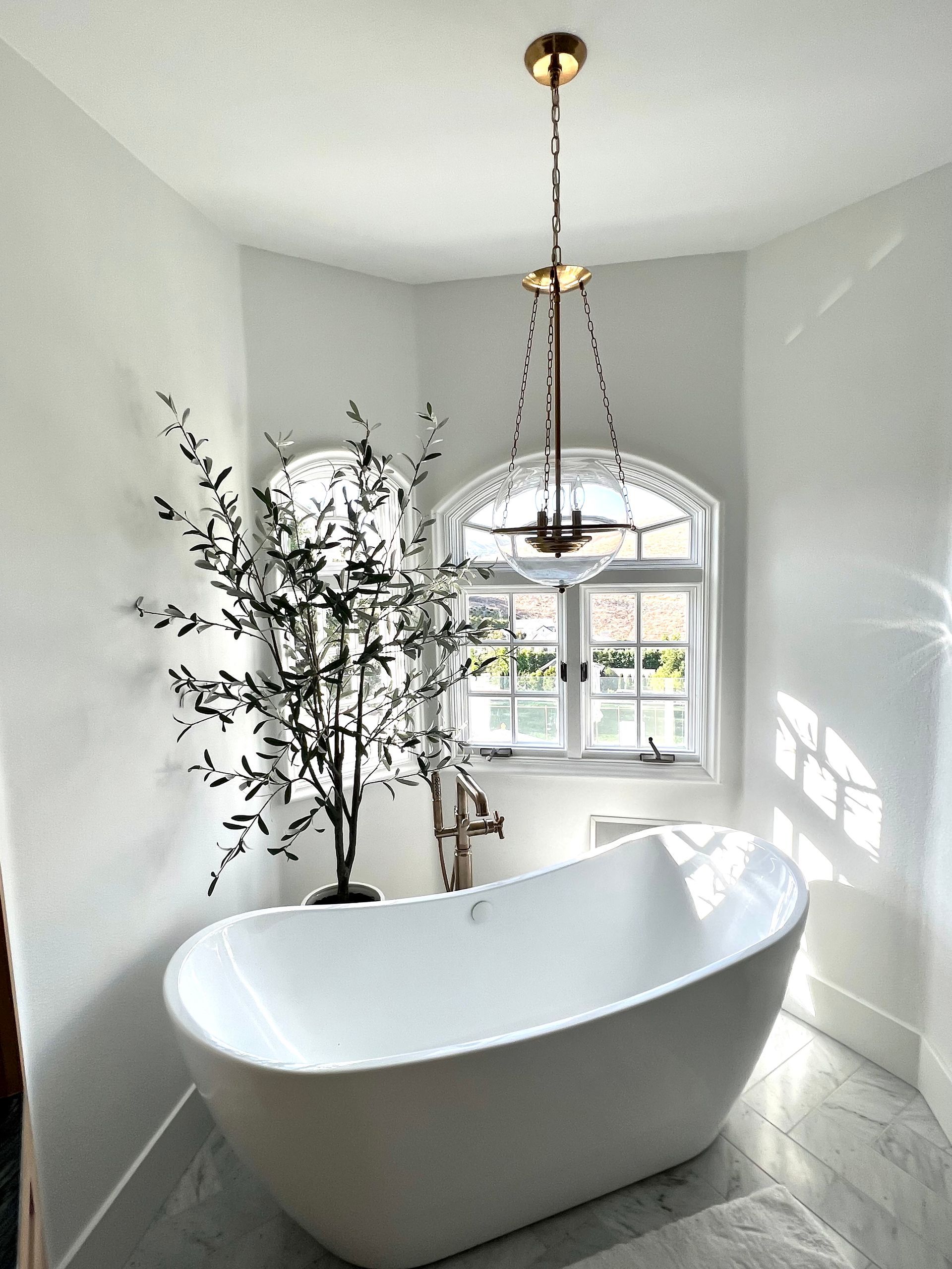 White bathtub in a bright, corner bathroom with a window and chandelier, and a black plant.