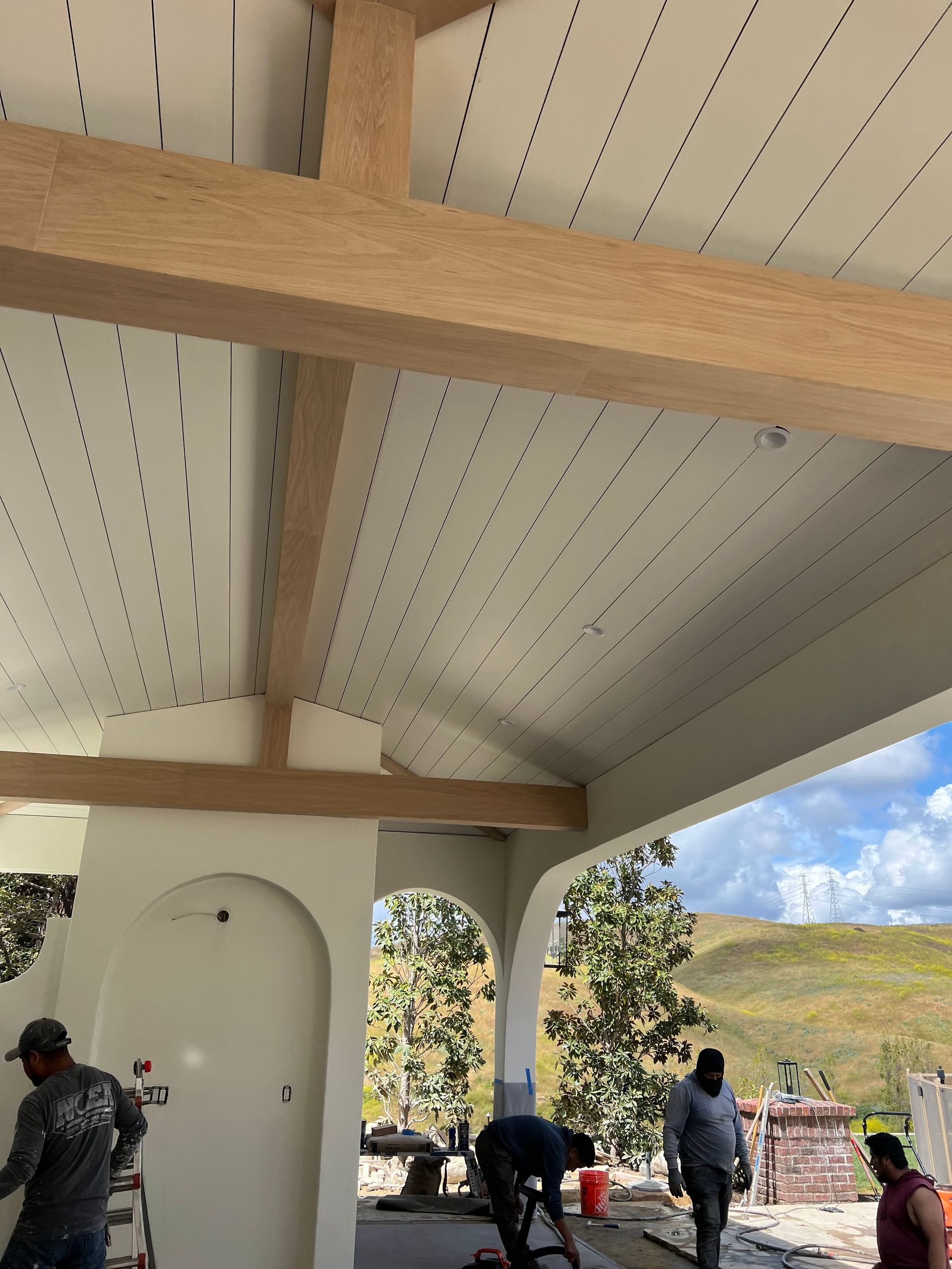 Workers installing a white perforated ceiling with wooden beams, under a white stucco overhang.