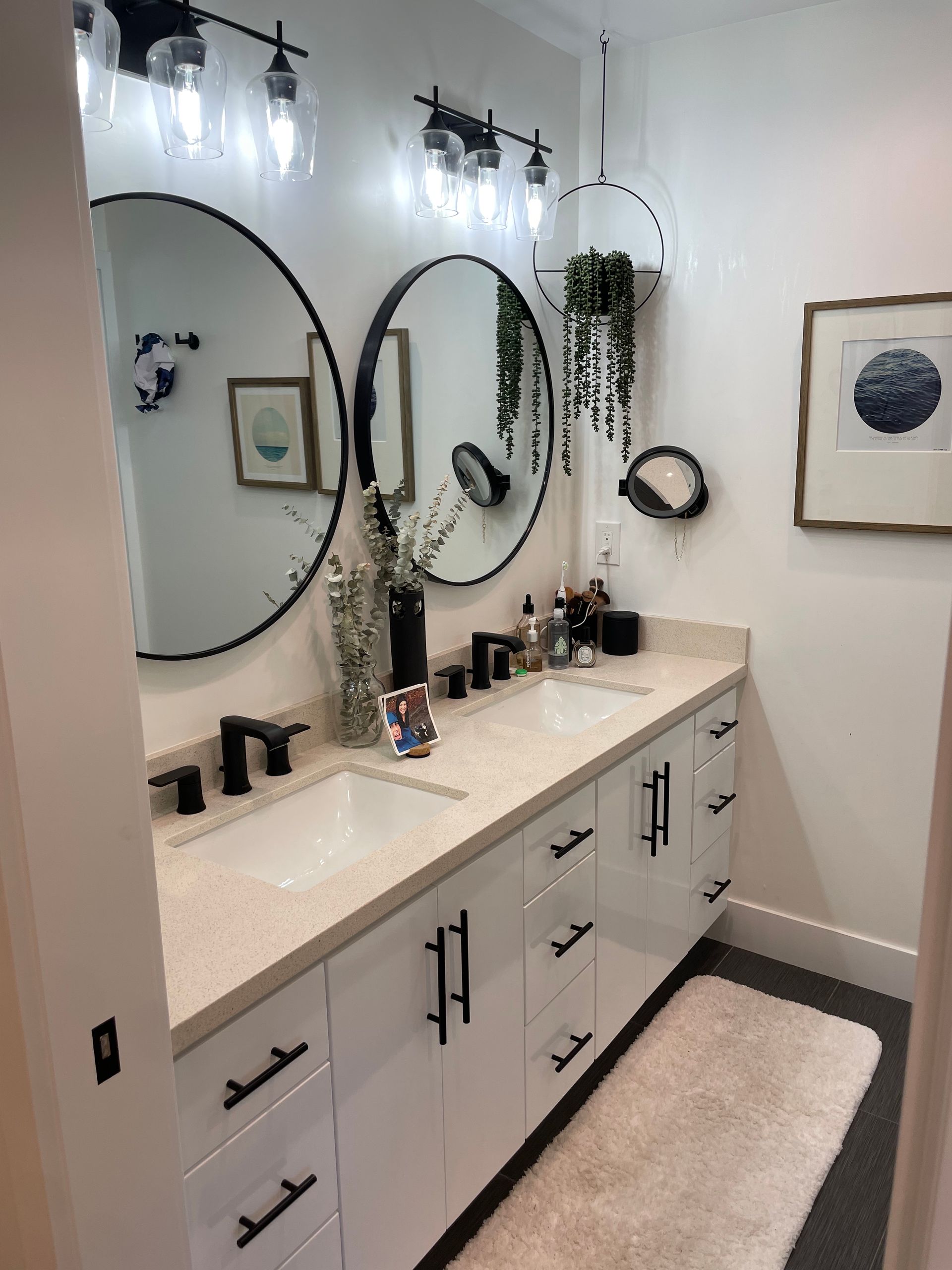Bathroom with white cabinets, two round mirrors, black fixtures, and a fluffy rug.