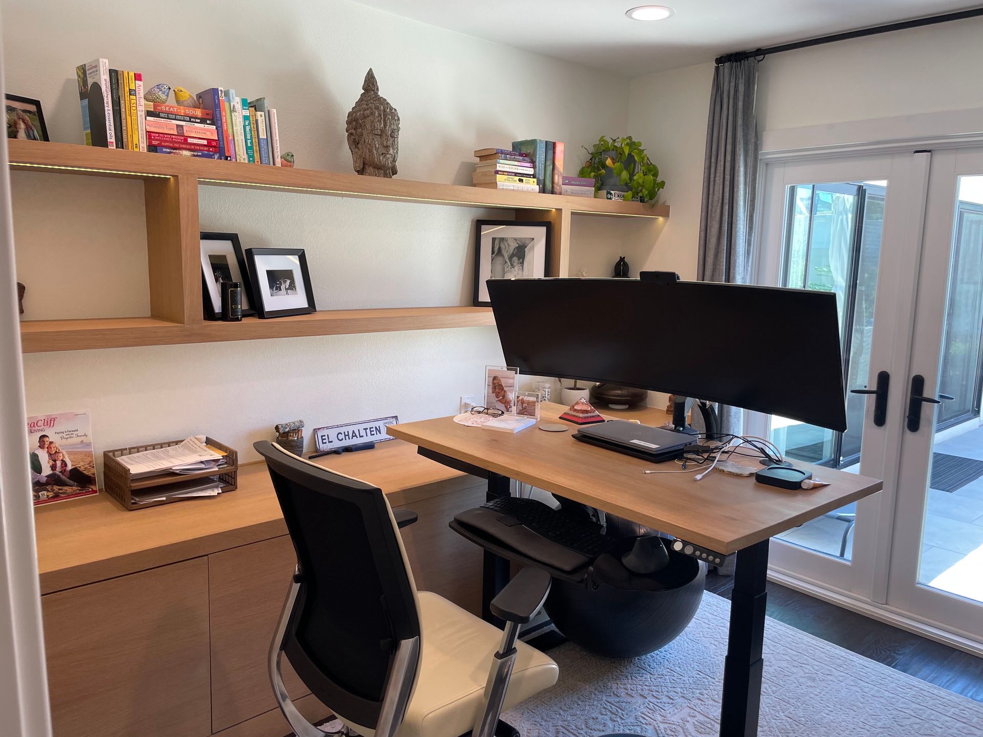 Office with a desk, shelves, and a large monitor. Light wood tones, neutral walls, and a door to the outside.
