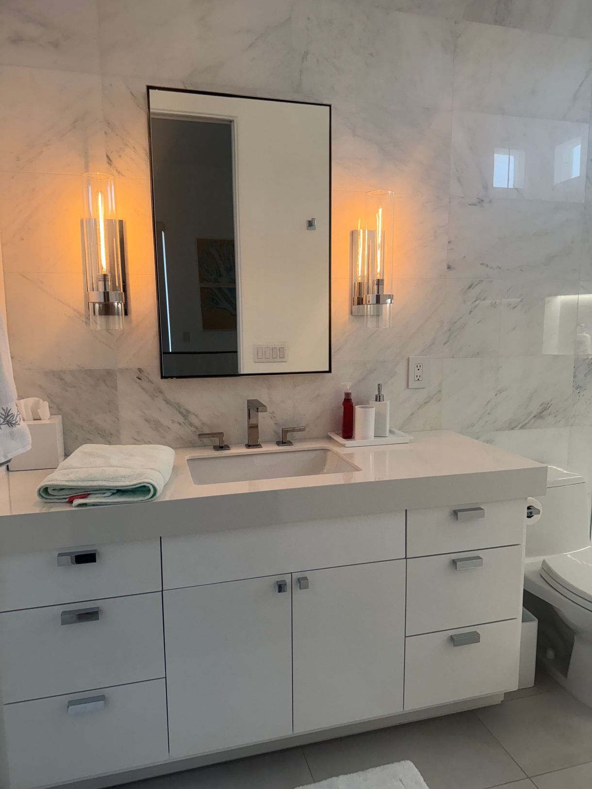 White bathroom vanity with marble wall, mirror, and sconces.