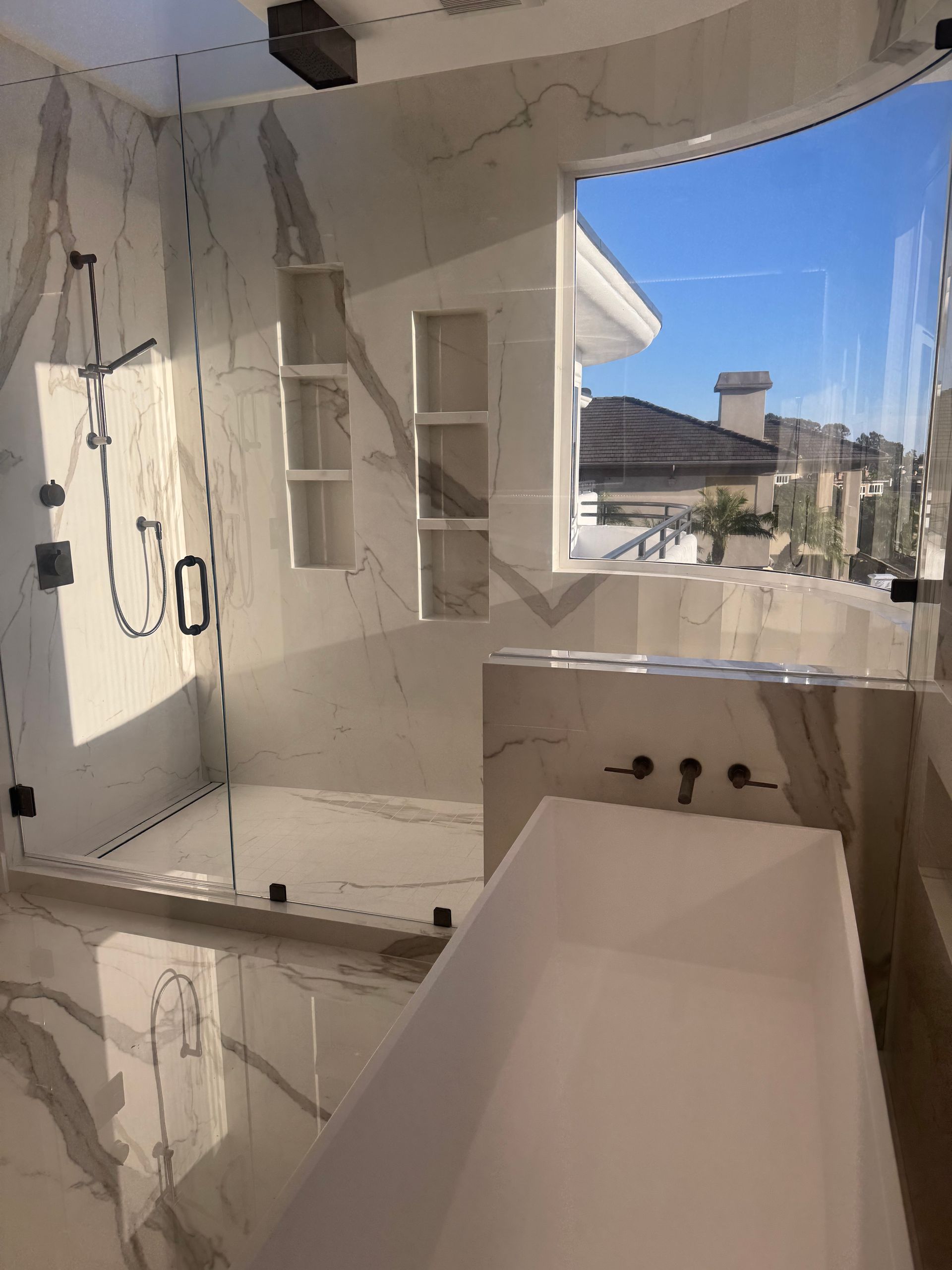 Luxurious bathroom with marble walls, glass shower, and freestanding tub. Window view of rooftops and sky.