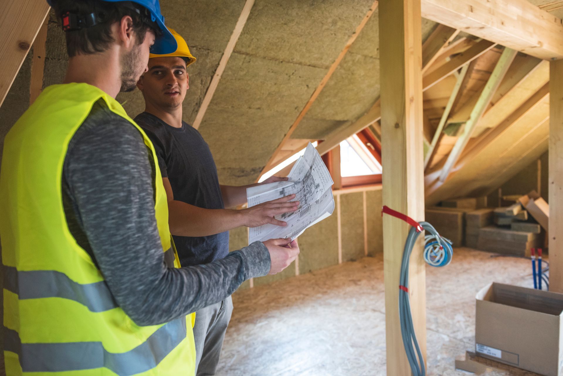 Two construction workers are looking at a blueprint in an attic.