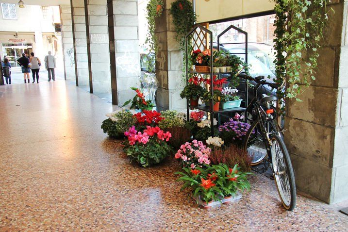 Fiori e biciclette esposti sotto un porticato; persone che camminano sullo sfondo.