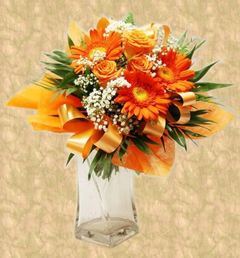Bouquet di margherite Gerber arancioni e rose in un vaso trasparente, legato con un nastro su uno sfondo decorato.