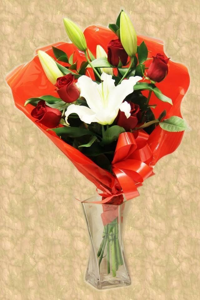 Bouquet di rose rosse e gigli bianchi, avvolti in carta rossa, in un vaso trasparente.