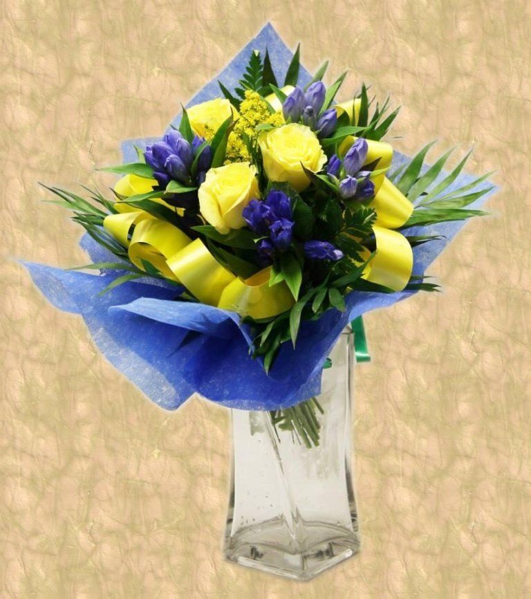 Bouquet di rose gialle e iris viola in un vaso trasparente, avvolto in carta velina blu e nastro.