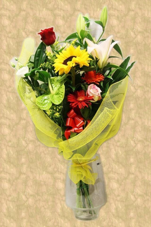 Mazzo di fiori colorati: rosa rossa, girasole, gigli, gerbera, avvolti in una rete gialla, in un vaso.