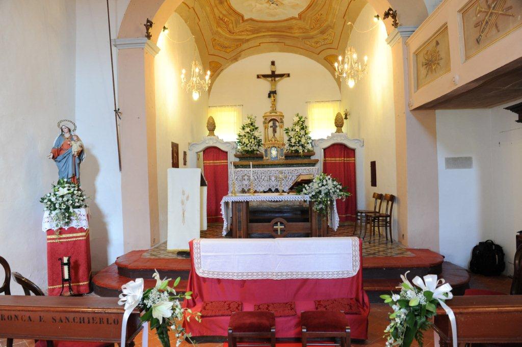 Interno di una chiesa. L'altare è decorato con fiori e un crocifisso. Sulla sinistra si trova una statua di Maria.