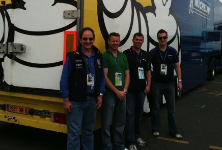 Four men standing in front of a Michelin truck. They wear lanyards and casual clothes, smiling.