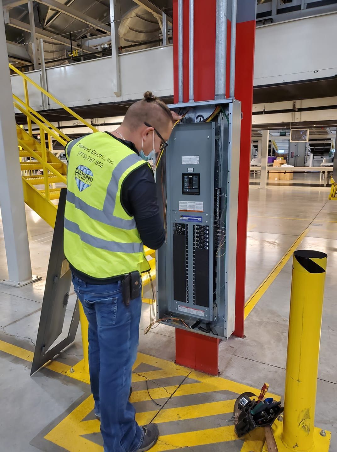 A man in a yellow vest is working on an electrical box in a warehouse.