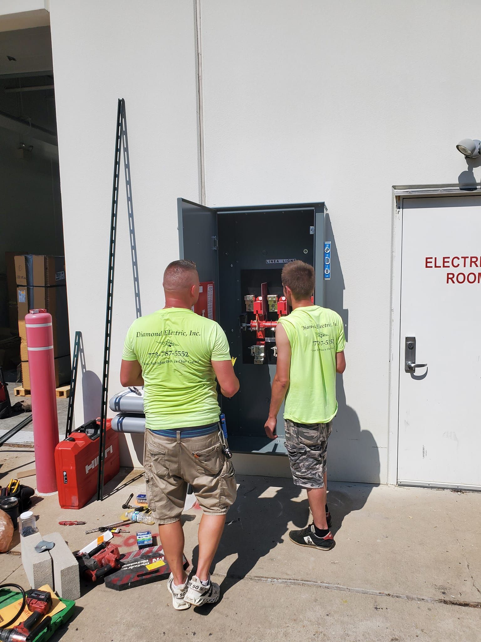 Two men in neon green shirts are standing in front of an electric box