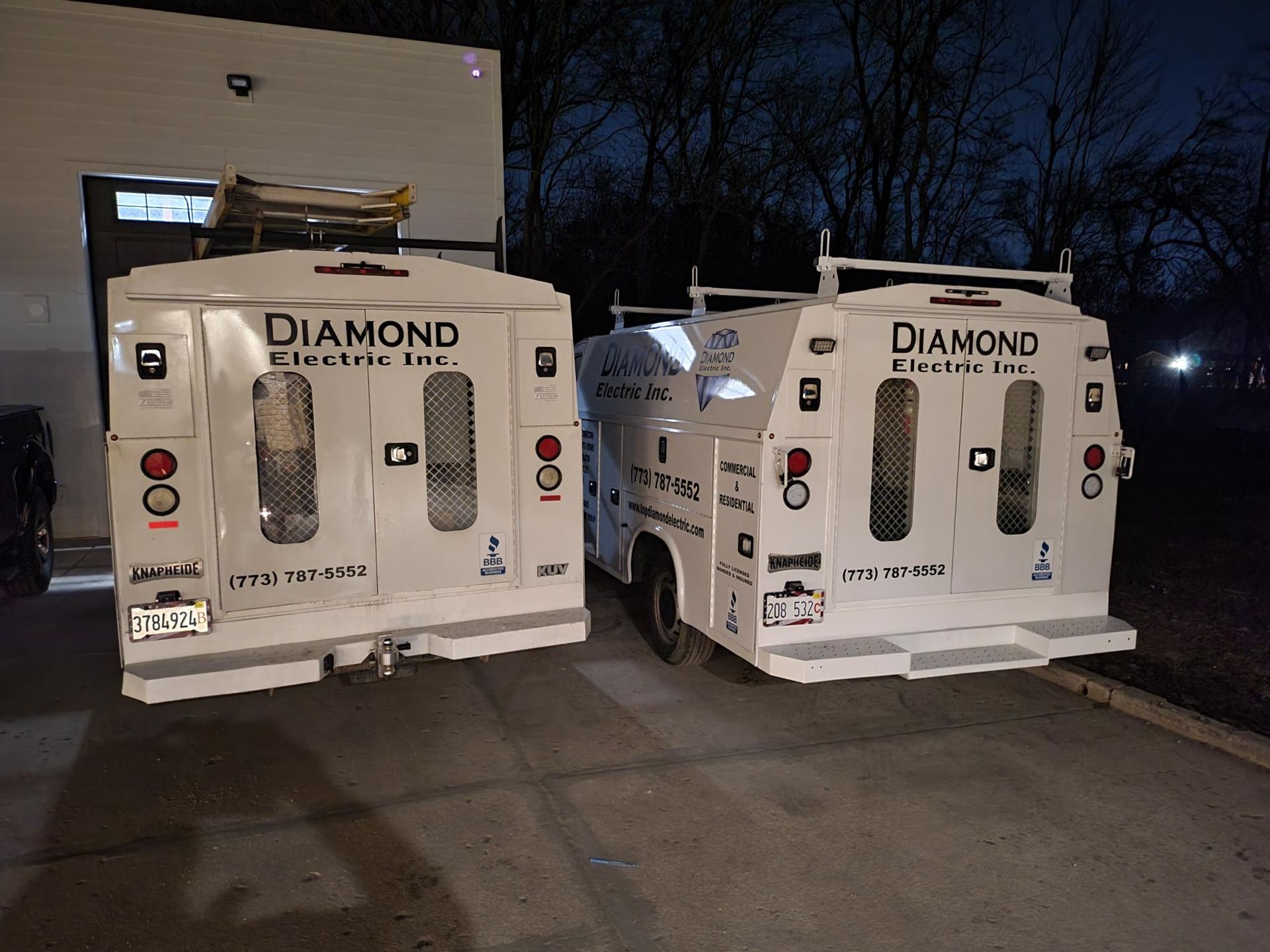 Two diamond electric trucks are parked in front of a building at night.