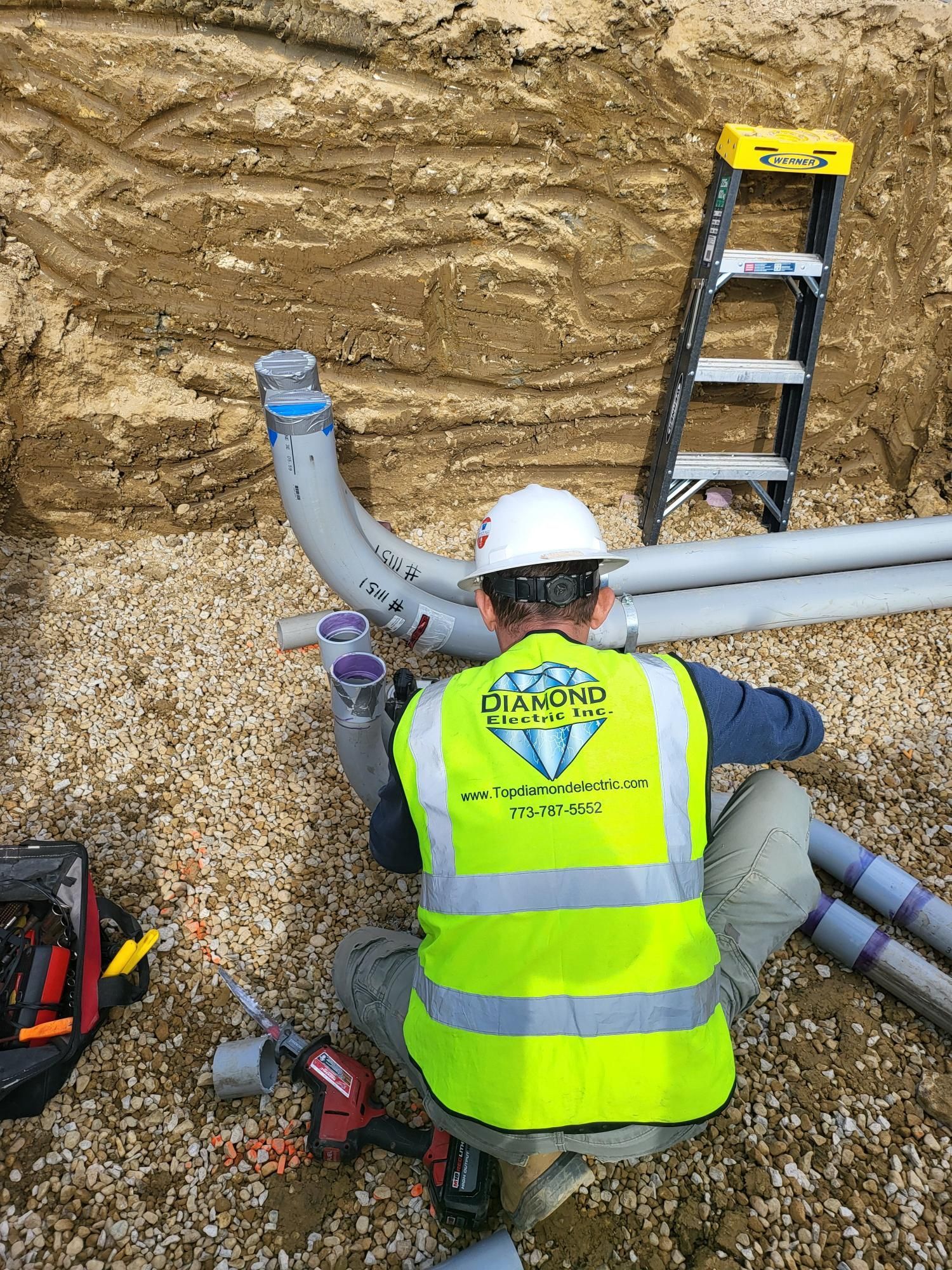 A man in a yellow vest and hard hat is working on pipes in the dirt.