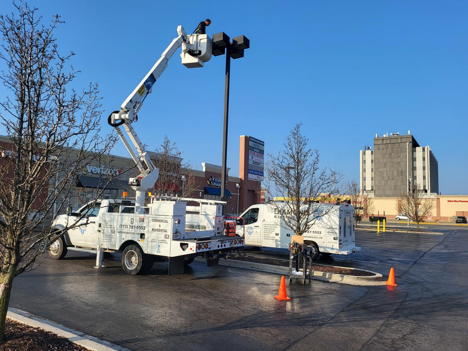 Two utility trucks are working on a street light in a parking lot.