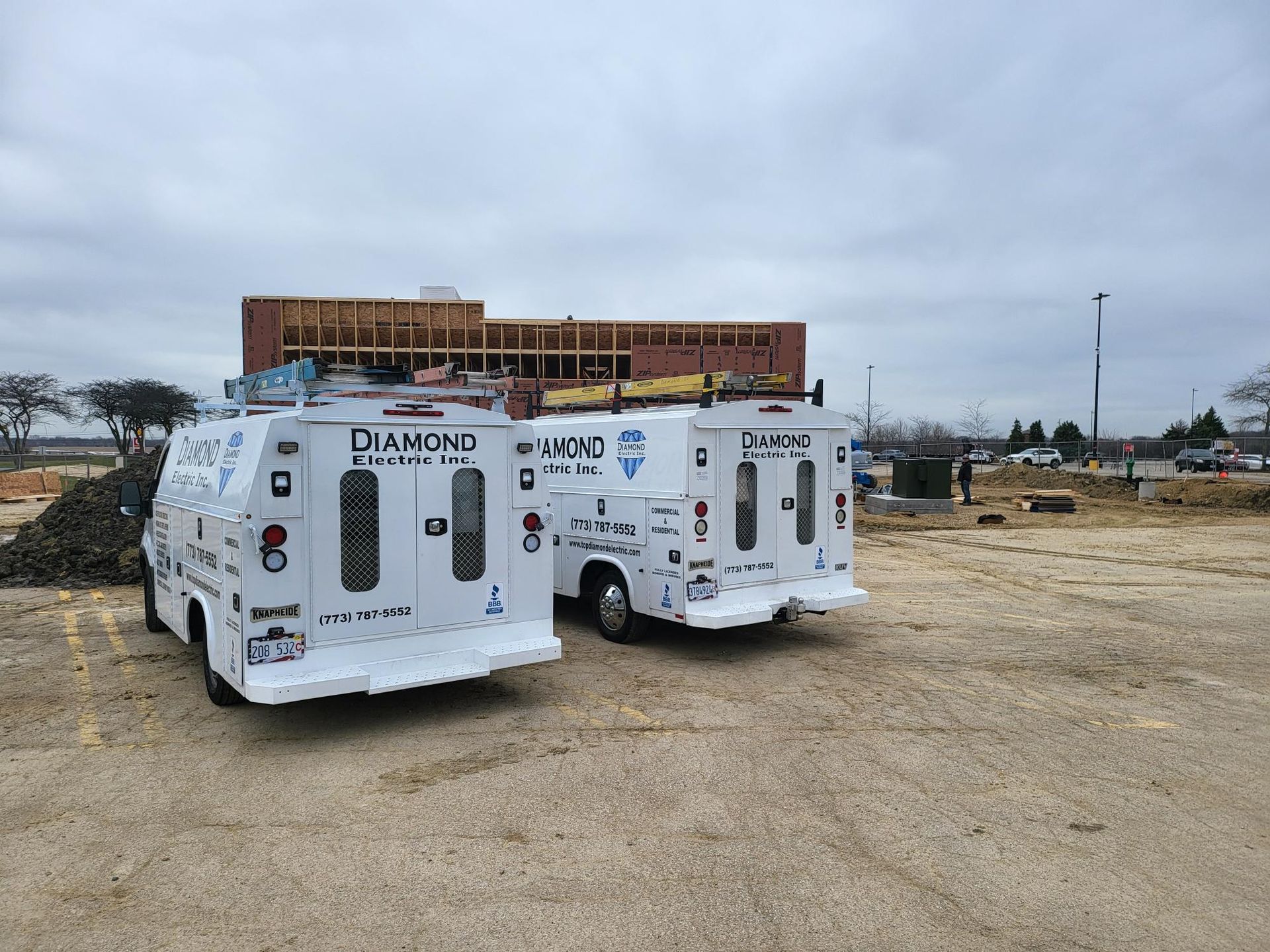 Two white vans are parked in a dirt lot in front of a building under construction.