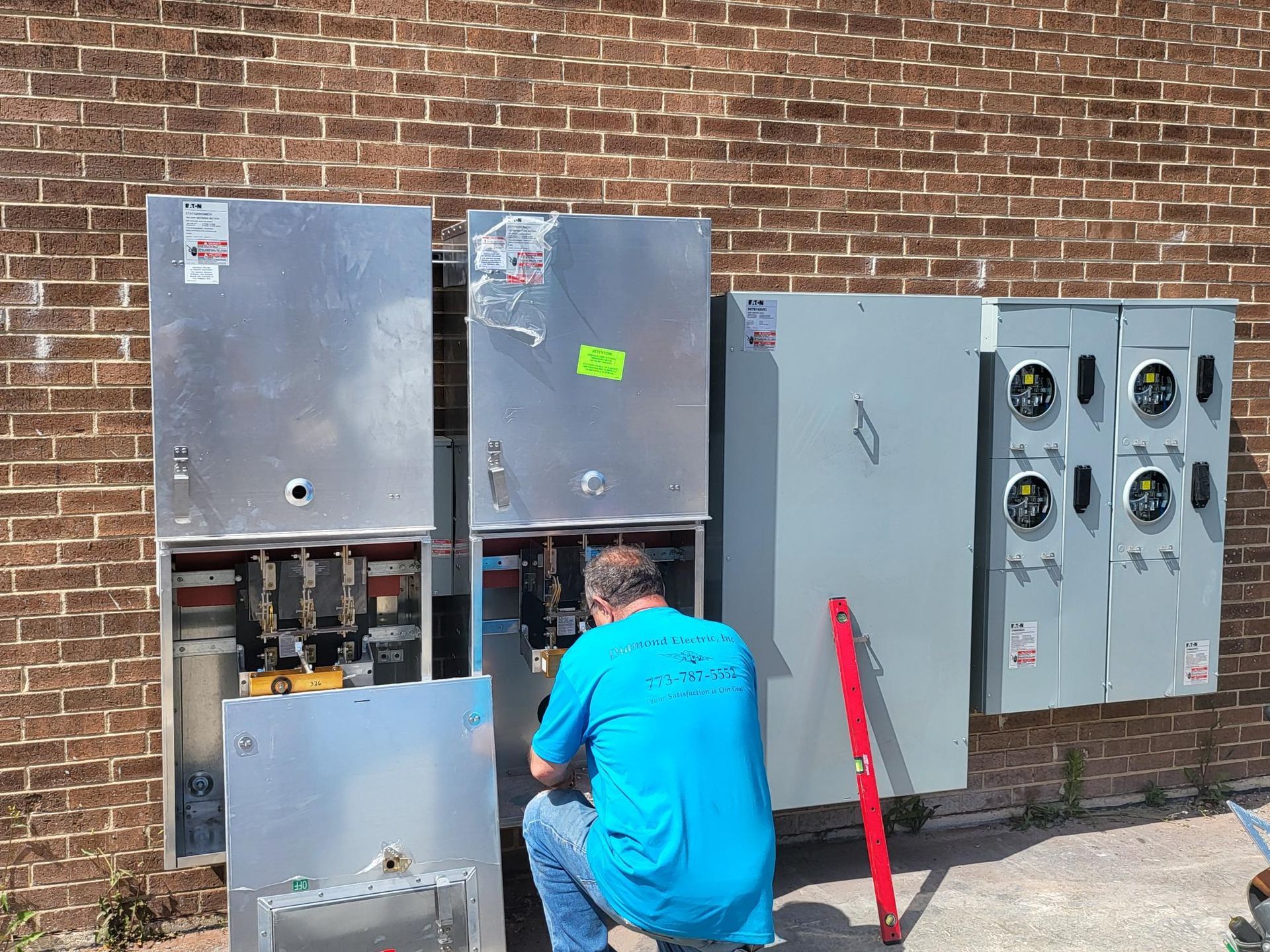 A man in a blue shirt is working on a electrical box on the side of a brick building.