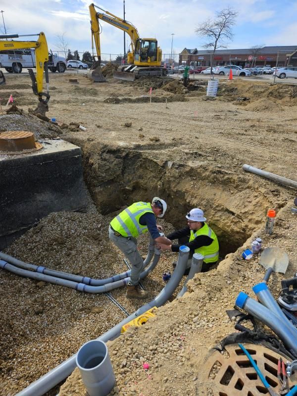 Two construction workers are working in a trench with a yellow excavator in the background.