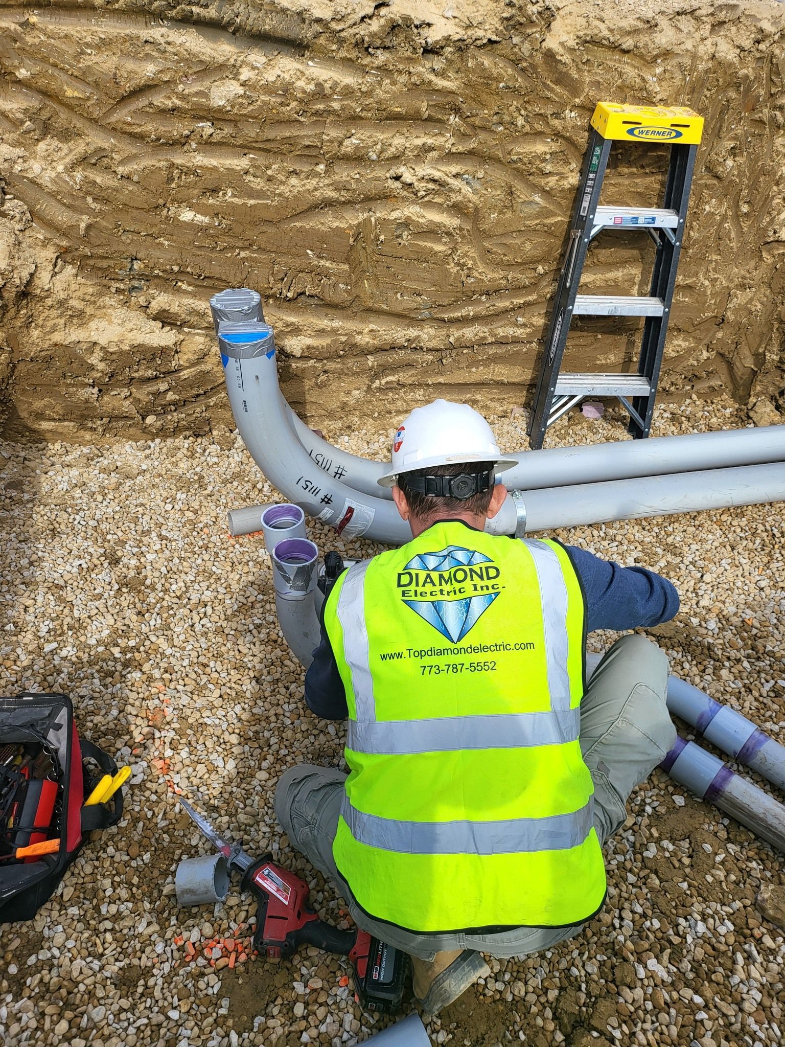 A man in a yellow vest is working on a pipe in the dirt.
