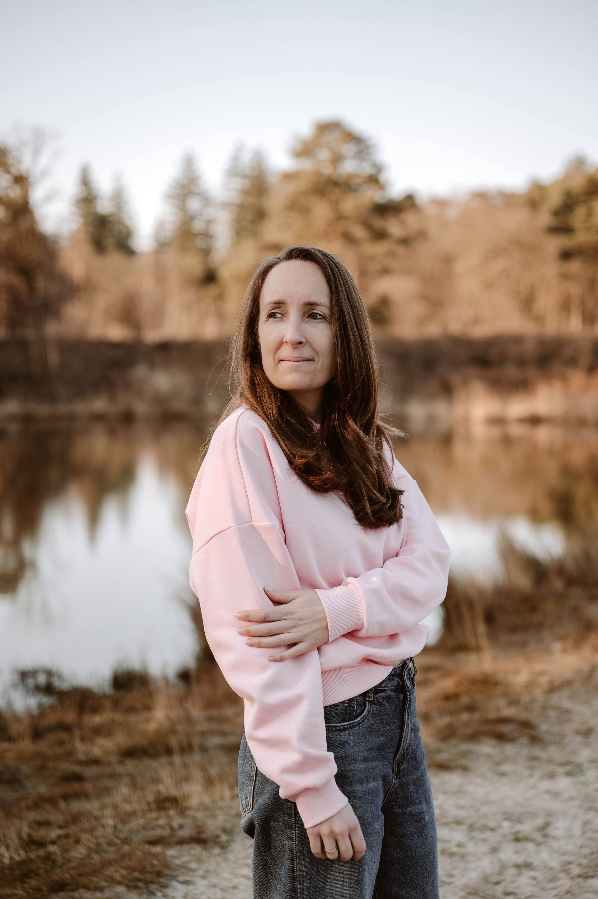 Vrouw in een roze trui staat bij een meer waar herfstbomen in het water weerspiegeld worden.
