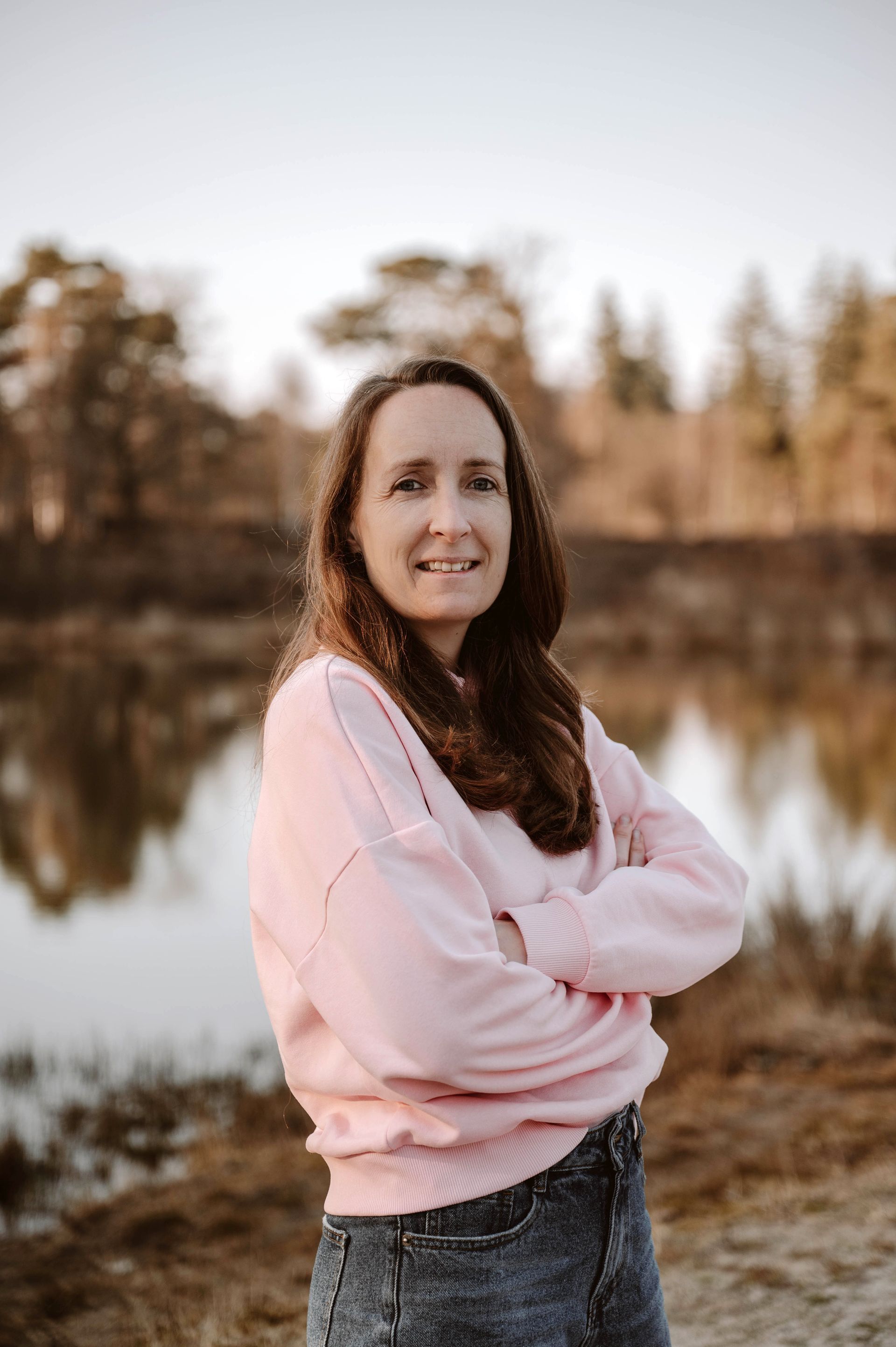 Vrouw in een roze trui staat buiten bij een spiegelend meer met herfstbomen op de achtergrond.