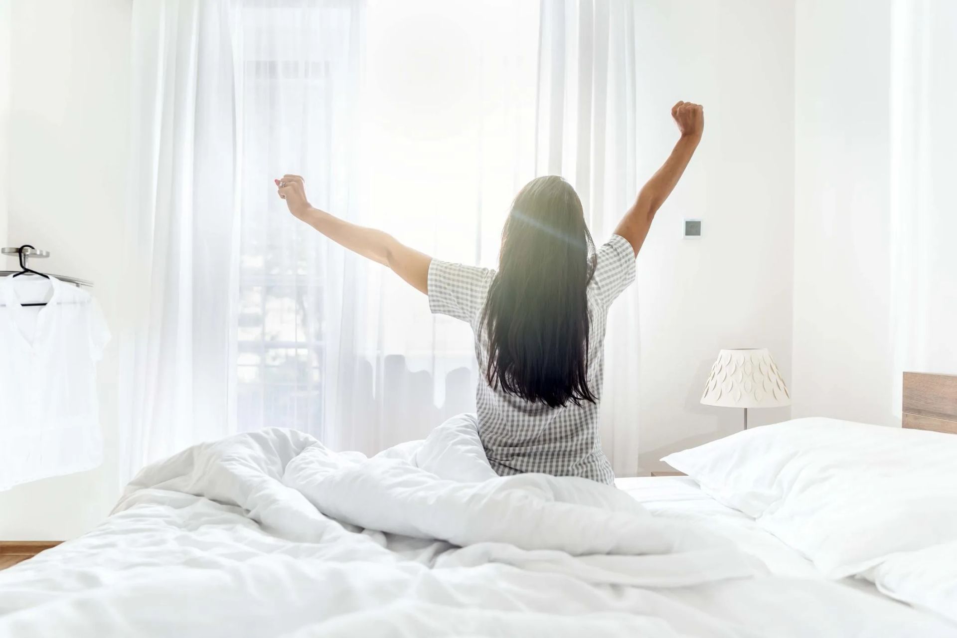 Een persoon rekt zich uit in bed in een lichte, witte slaapkamer met doorschijnende gordijnen en zonlicht.
