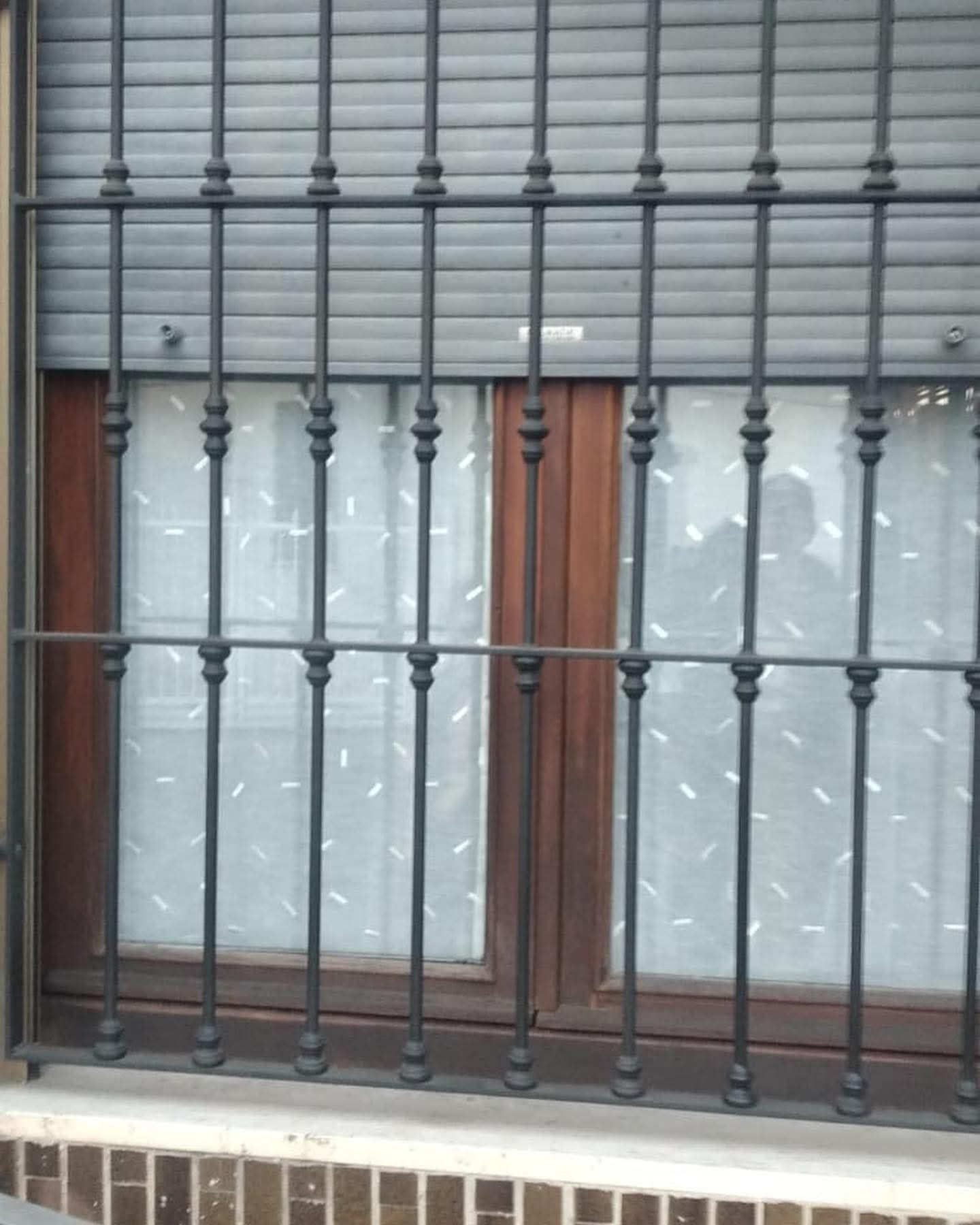 Finestra con sbarre di sicurezza in ferro battuto, telaio in legno e tende bianche a fantasia.