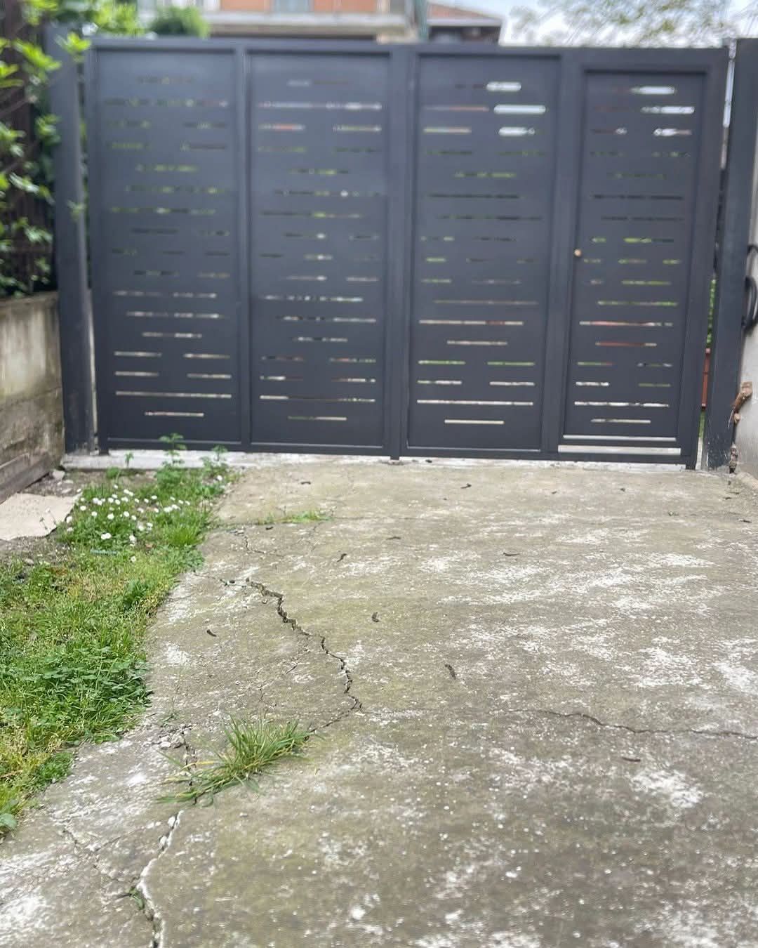 Cancello in metallo grigio con ritagli orizzontali, su vialetto in cemento con erbacce.
